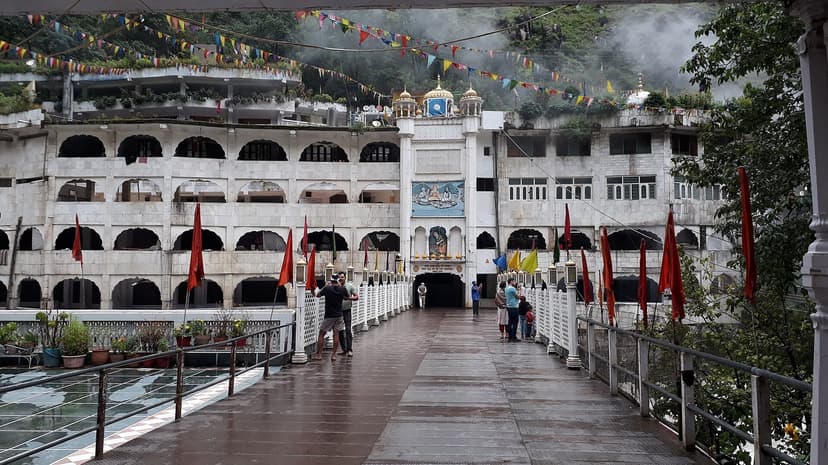 White arched Manikaran Gurudwara complex with prayer flags, pilgrims on the walkway, steam from hot springs rising behind