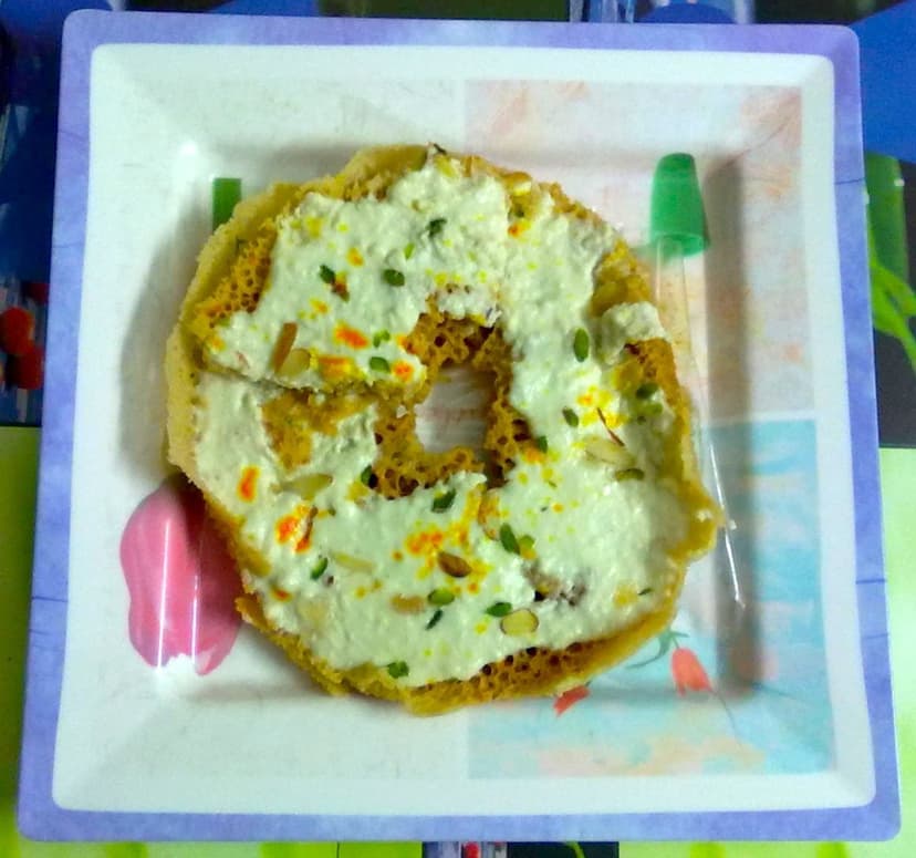 A round ghevar, a honeycomb-textured Rajasthani sweet, topped with a thick layer of white rabri cream, chopped pistachios and saffron, on a plate