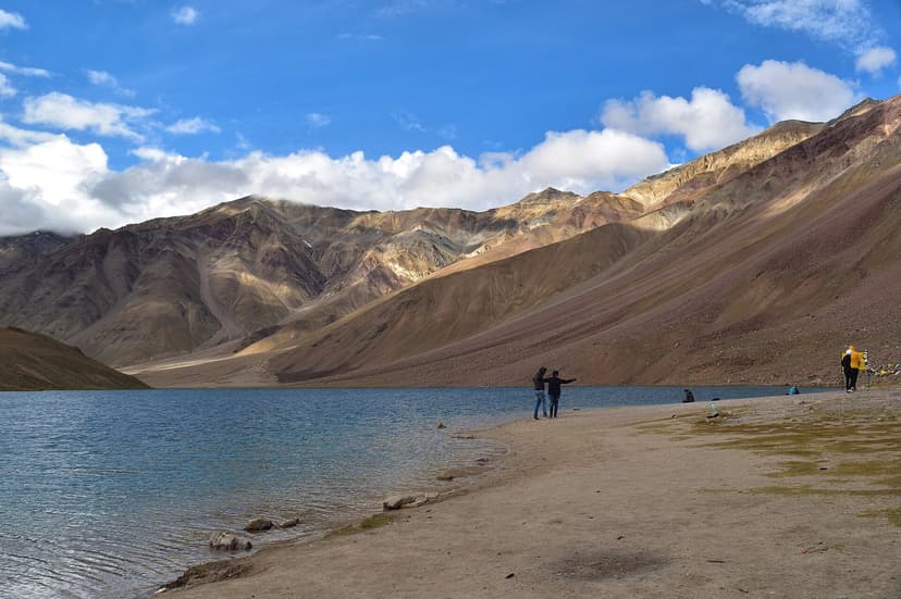 Chandratal Lake at 4,300 m in Lahaul-Spiti, turquoise water lapping the gravel beach, rust-and-green ridges of the Chandra Bhaga ranges behind, two figures by the shore for scale