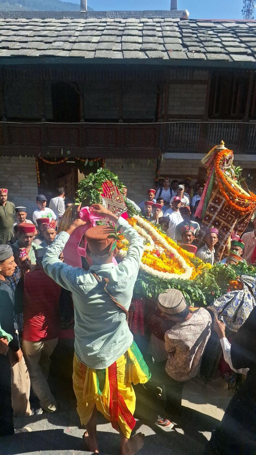 A village deity in a garlanded palanquin carried by men in Kullu caps past a wooden kath-kuni house