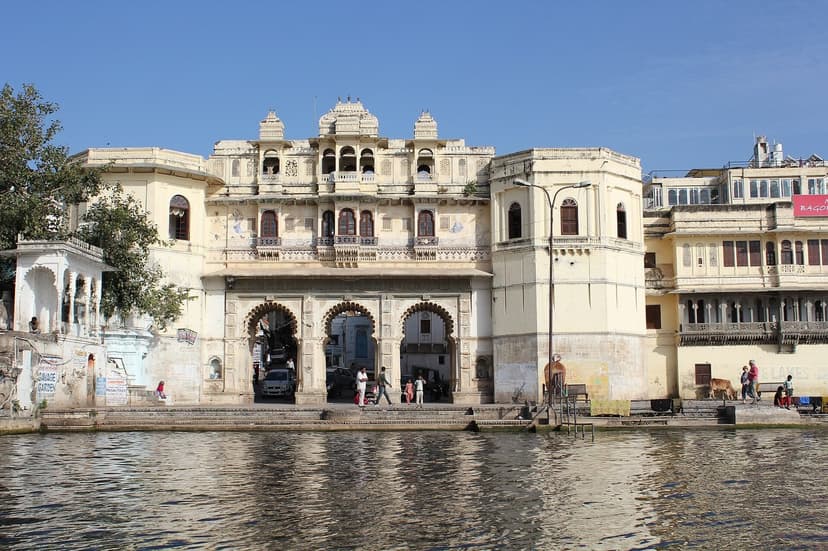A cream heritage haveli with arched gateways rising from the water at Lal Ghat on Lake Pichola, Udaipur, a few people and a cow on the ghat in front.
