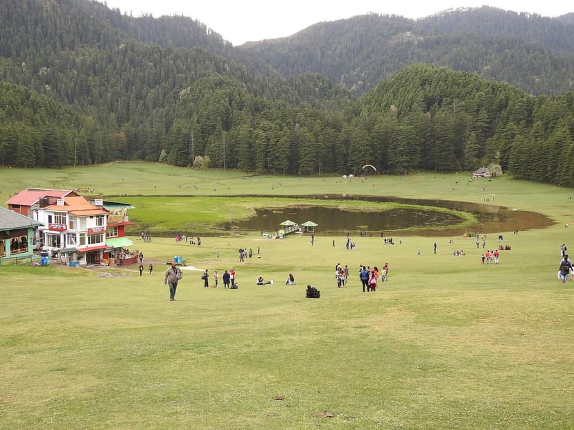 The saucer-shaped Khajjiar meadow in Chamba district, wide green grass plain with a small lake in the centre, ringed by a thick wall of deodar cedar forest, with people walking and sitting on the grass