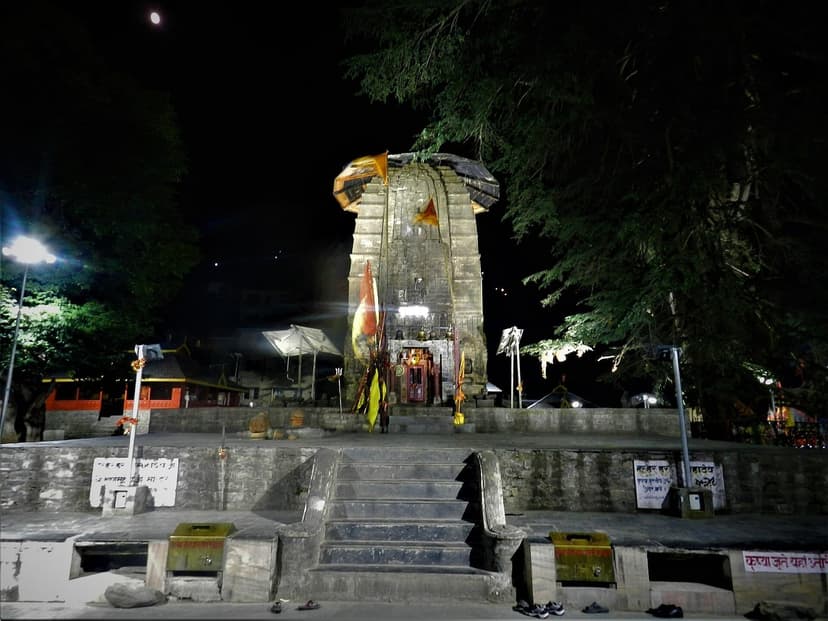 Manimahesh Temple of Bharmour lit at night, moon visible above the Shikhara, with a few pairs of shoes left at the steps and devotional flags hanging at the entrance