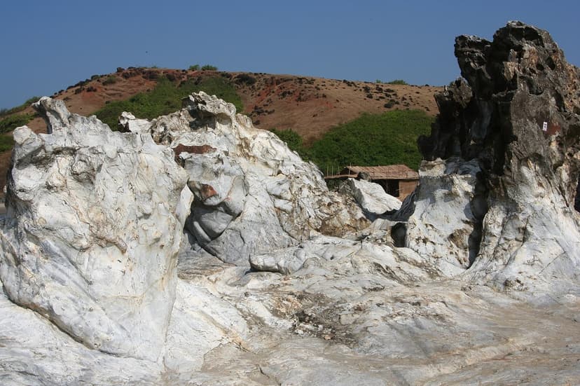Large pale weathered rock formations on the shore at Vagator, Goa, with a scrubby red-brown headland and a hut behind under a clear blue sky.