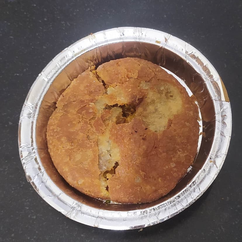A single golden-brown pyaz kachori in a foil bowl, split open to show the spiced onion filling