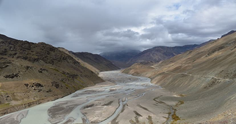 Wide panoramic view of the Chandra River braided across a flat grey gravel valley floor, brooding low cloud overhead, brown ridges either side, the road visible as a thin line on the right edge
