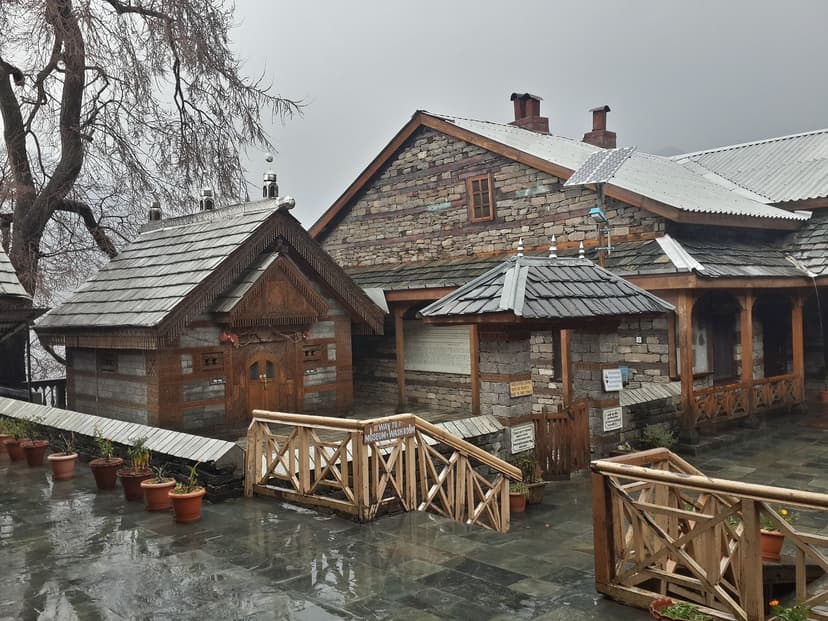 Naggar Castle in Kullu district photographed on a rainy day, centuries-old kath-kuni stone and deodar wood construction with pitched slate roofs, wooden balconies and a courtyard in the foreground