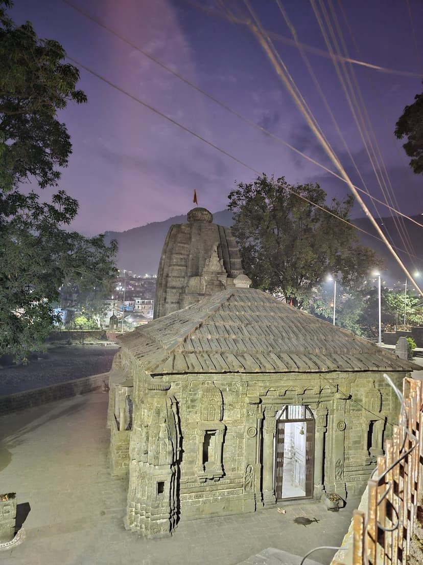 The stone shikhara of Triloknath Temple in Mandi at twilight, with a tiled mandap in front and town lights behind