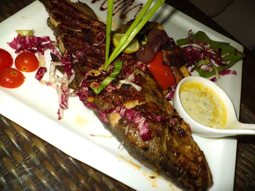 Whole grilled Himalayan trout served on a white plate with red cabbage slaw, cherry tomatoes and a side of butter sauce, the river fish that defines Kullu valley non-veg