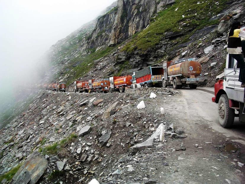 A long queue of cargo trucks stopped on the rubble-strewn Rohtang road with fog filling the gorge below and loose rock above