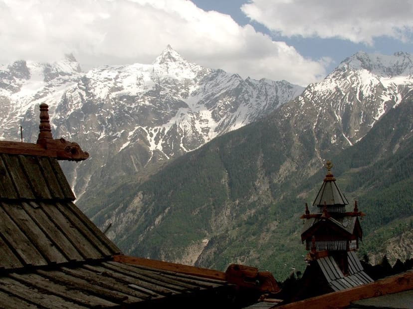 Wood-shingle Kinnauri rooftops and a temple spire in Kalpa with the snow peaks of the Kinner Kailash range behind, mid-day cloud