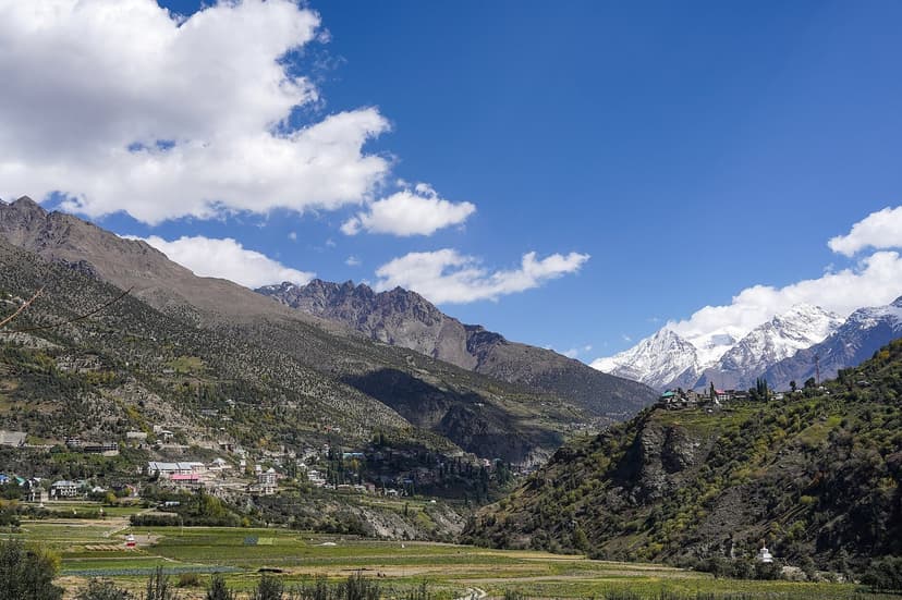 Wide view of Keylong town in the Lahaul valley, a cluster of buildings on a green slope above the river, with snow-capped peaks rising behind and barley fields in the foreground