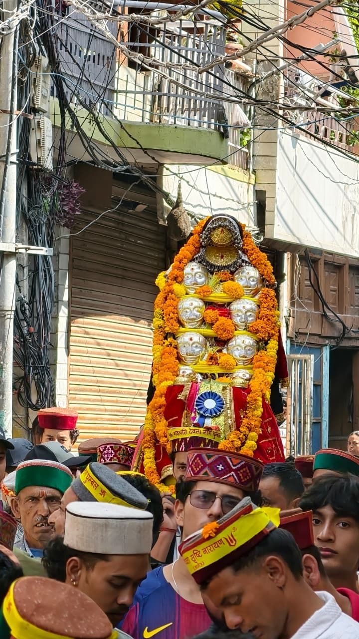 A deity palanquin covered in silver mohras and marigold moves through a Kullu street with a crowd