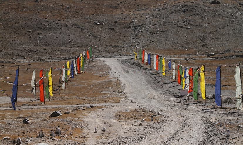 Brown gravel track climbing toward Kunzum La pass at 4,590 m, lined on both sides by tall poles draped with multicolour Tibetan prayer flags. Barren mountain bowl in background
