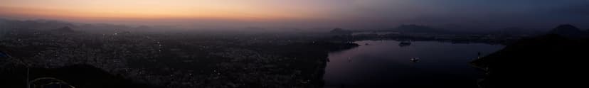 Wide dawn panorama over Udaipur city and Fateh Sagar Lake seen from the Neemach Mata ridge, warm orange sky over silhouetted Aravalli hills.