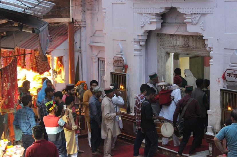 A Himachali devta procession entering a temple gate with men in traditional Kullu caps carrying the deity palanquin and beating drums while onlookers crowd around