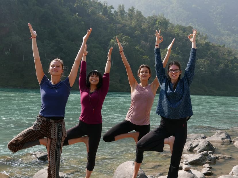 Four people holding tree pose on rocks beside the turquoise Ganga at Rishikesh, forested hills behind, during a yoga teacher training