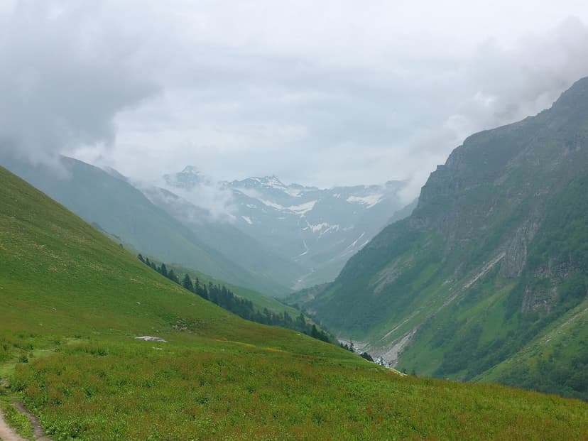 A wide green high-altitude meadow valley with mist, snow patches and a stream in the Western Himalayas