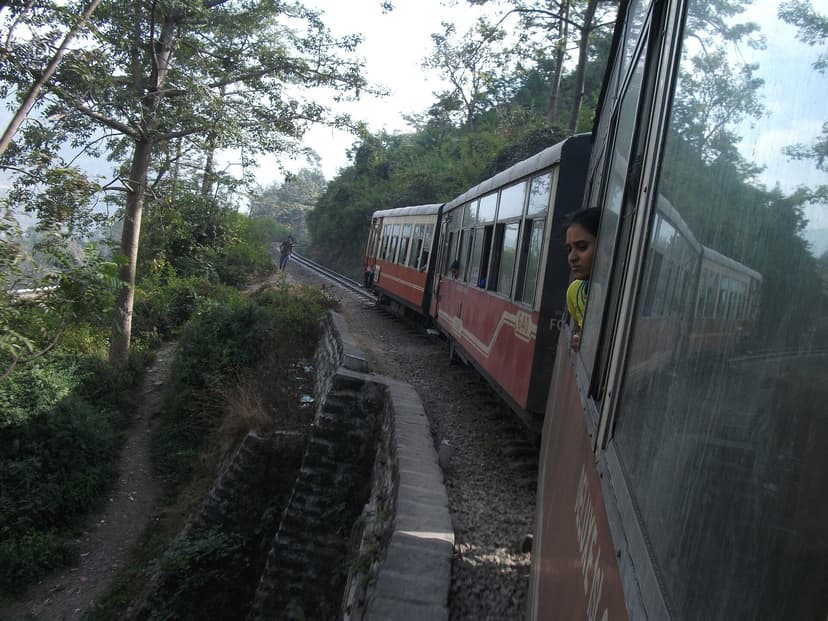 Red and cream Kalka-Shimla narrow gauge toy train curving through green hillside with pine trees