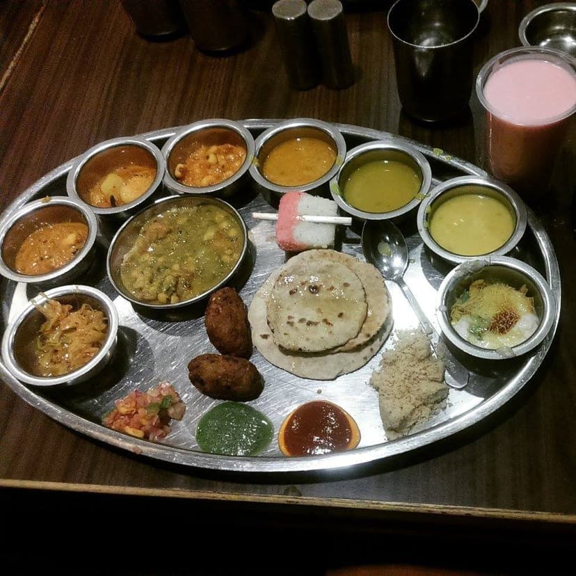 A Rajasthani thali on a steel plate with many small bowls of curries, dal, gatte, kadhi, chutneys and papad, baati and roti in the centre, churma, and a glass of pink lassi