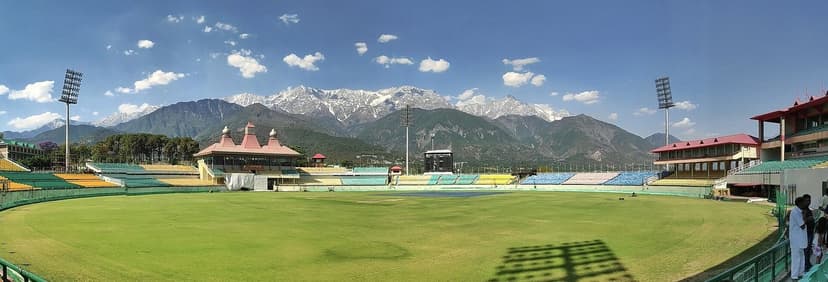 The HPCA Cricket Stadium in Dharamshala shot from the boundary line, colourful empty stands in yellow green and blue, a small Himachali-style pavilion at the midwicket end, the Dhauladhar range fully snow-capped behind, a few staff on the outfield