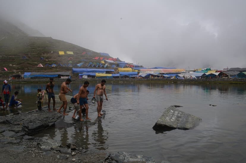 Pilgrims bathing at Manimahesh Lake in monsoon mist with colourful tarpaulin tent village ringing the shore