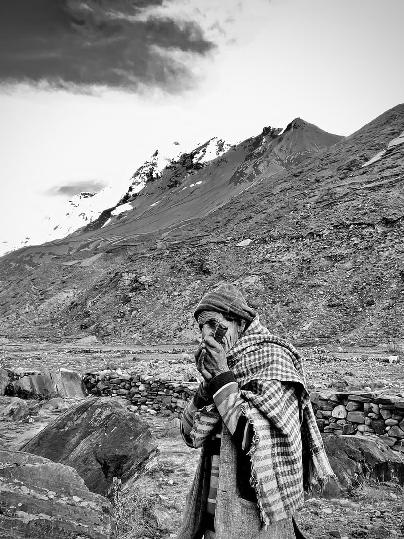 Black and white portrait of an elderly Gaddi shepherd at rest, wearing a chequered gardu blanket and a topi, holding a pipe, with a snow-streaked ridge rising behind him