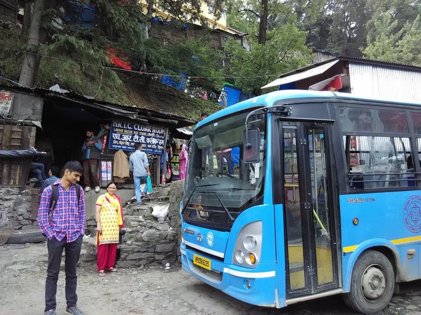 A blue HRTC deluxe bus parked at Lakkar Bazar bus stand in Shimla with hillside shops behind