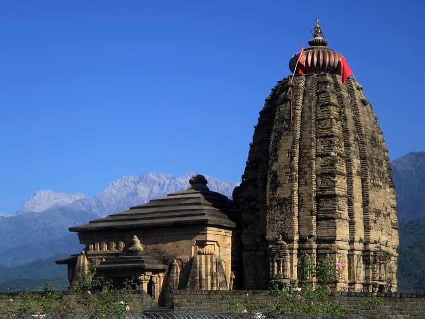 Baijnath Temple's carved stone shikhara from 1204 AD with a red flag on top and snow-covered Dhauladhar peaks behind, Kangra district