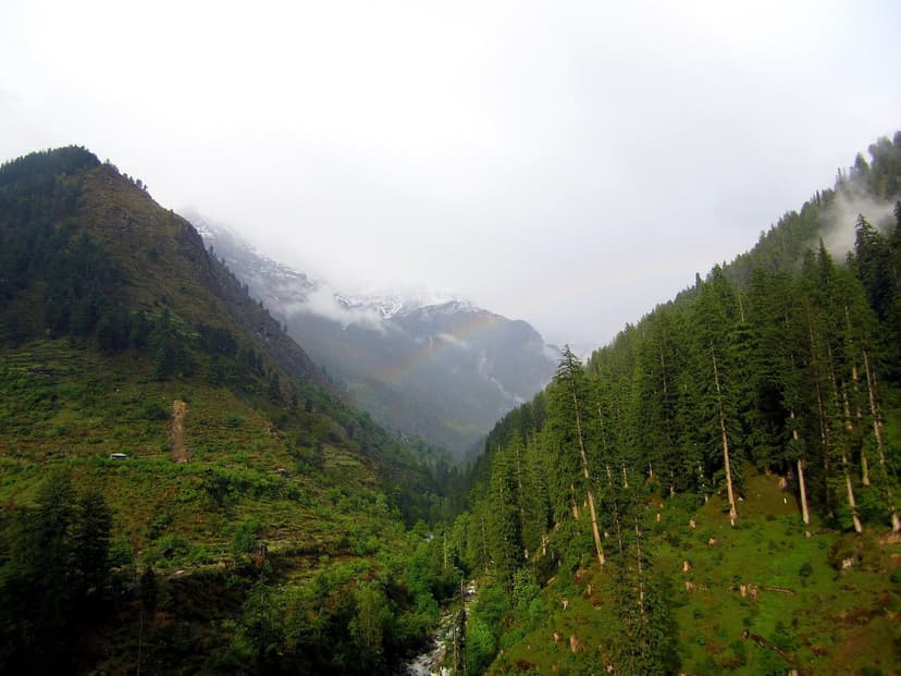 Cloud-hugged Parvati valley with a rainbow arcing across the slope, deodar forest, river trickle below