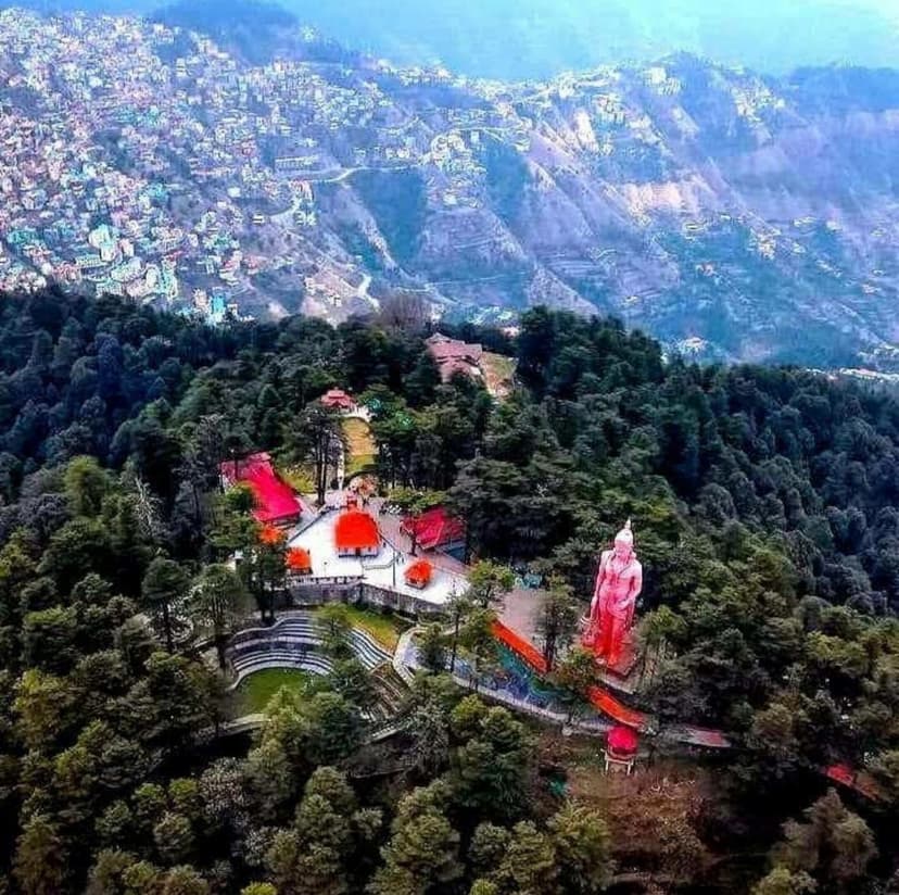 Aerial view of Jakhoo Temple with 108-foot red Hanuman statue on a forested hilltop, Shimla city visible in the background below