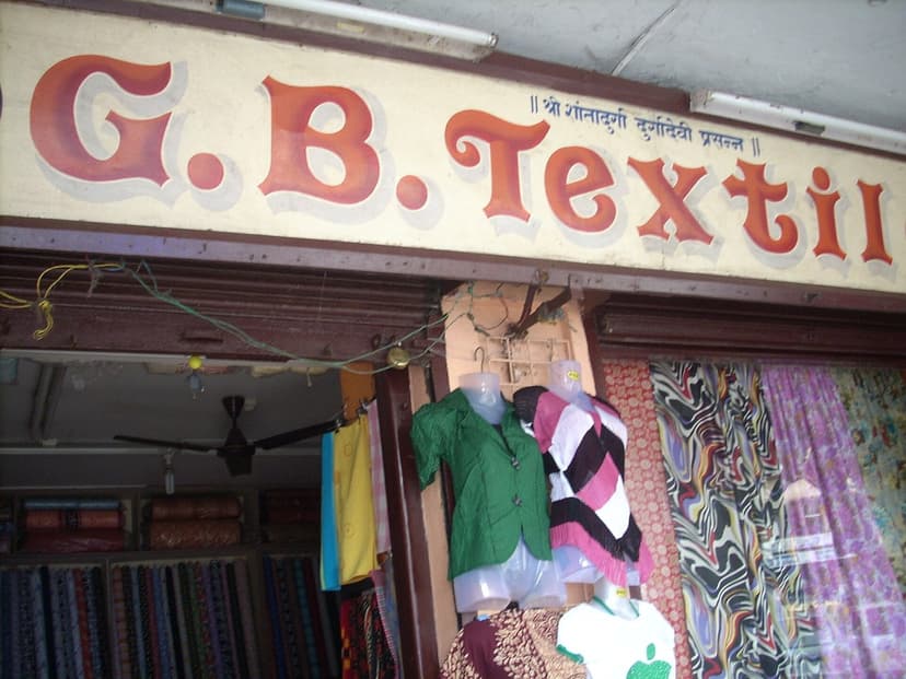 A hand-painted signboard reading G. B. Textiles above a fabric shop in Mapusa market, bolts of cloth and garments on display, no people