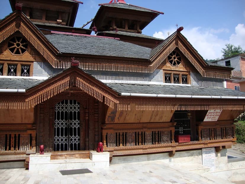 Kamaksha Devi temple at Kao near Karsog, a tiered slate-roofed kath-kuni pagoda with an ornately carved wooden facade and two small red stone lions at the door