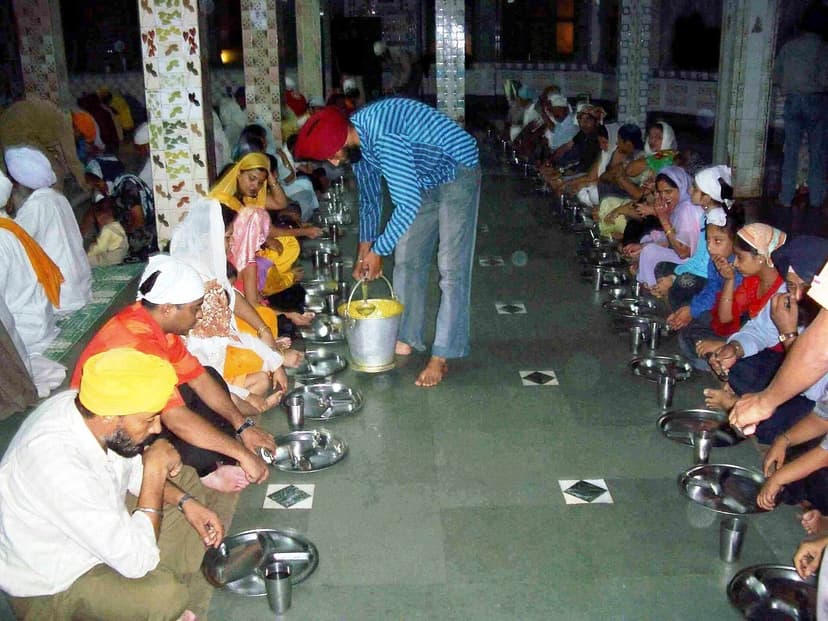 Two parallel rows of seated diners on a polished stone floor at the Sikh Gurdwara langar in Manikaran, Himachal Pradesh, with a server walking between rows pouring food from a steel bucket — structurally similar to the Painth seating used at Boti-prepared Dham feasts in Himachali villages