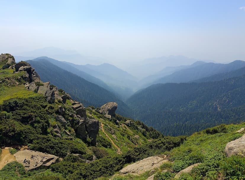 View from near Churdhar peak across granite outcrops and green meadow ridges, with hazy blue forested valleys of the Tons catchment receding below