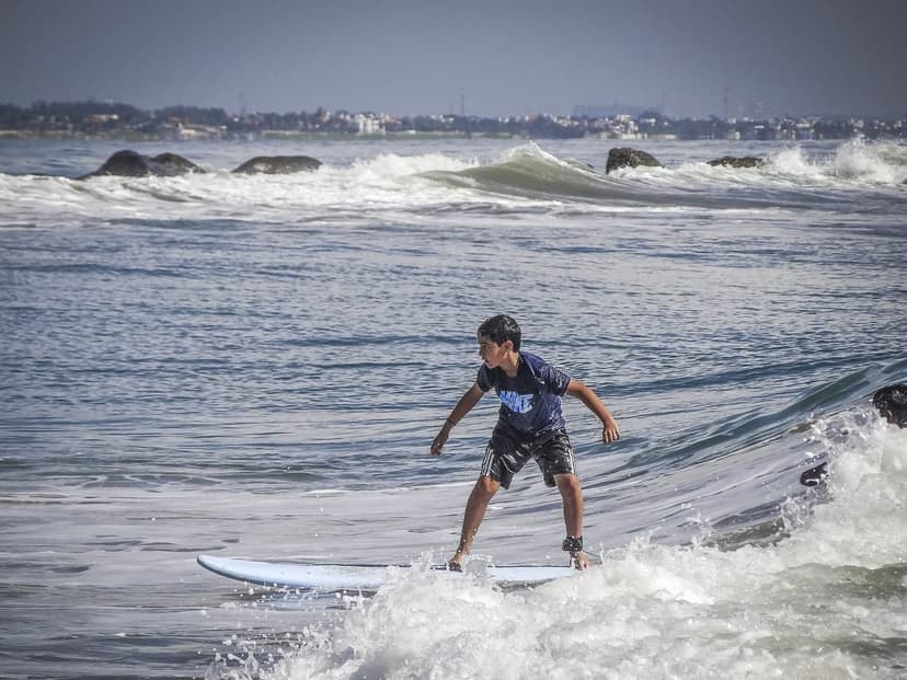 A young beginner surfer riding a light blue soft-top foam board across a small breaking wave, arms out for balance