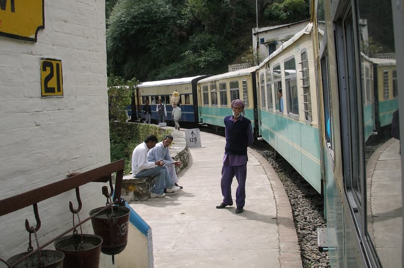 The narrow-gauge Kalka–Shimla heritage train at a small station with passengers and a porter on the platform, the green and cream carriages stretching down the line