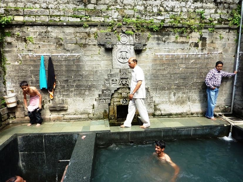 Stone-walled bathing pool at Vashisht hot springs near Manali with carved spout, three men present including one in the pool and two on the side, sulphurous water visible