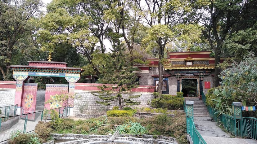 Tibetan-style red and white painted gateway with stupa visible inside the Norbulingka Institute grounds, set among trees outside Dharamshala
