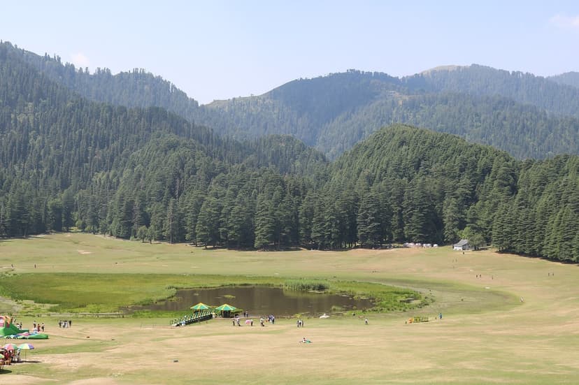 Wide view of Khajjiar meadow with central pond, families and horse handlers visible, ringed by tall deodar and oak forest with low Dhauladhar foothills in the background