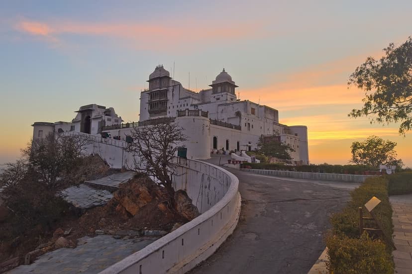 The white Sajjangarh Monsoon Palace on its hilltop at dusk near Udaipur, a winding road leading up to it under a pink and orange sky.