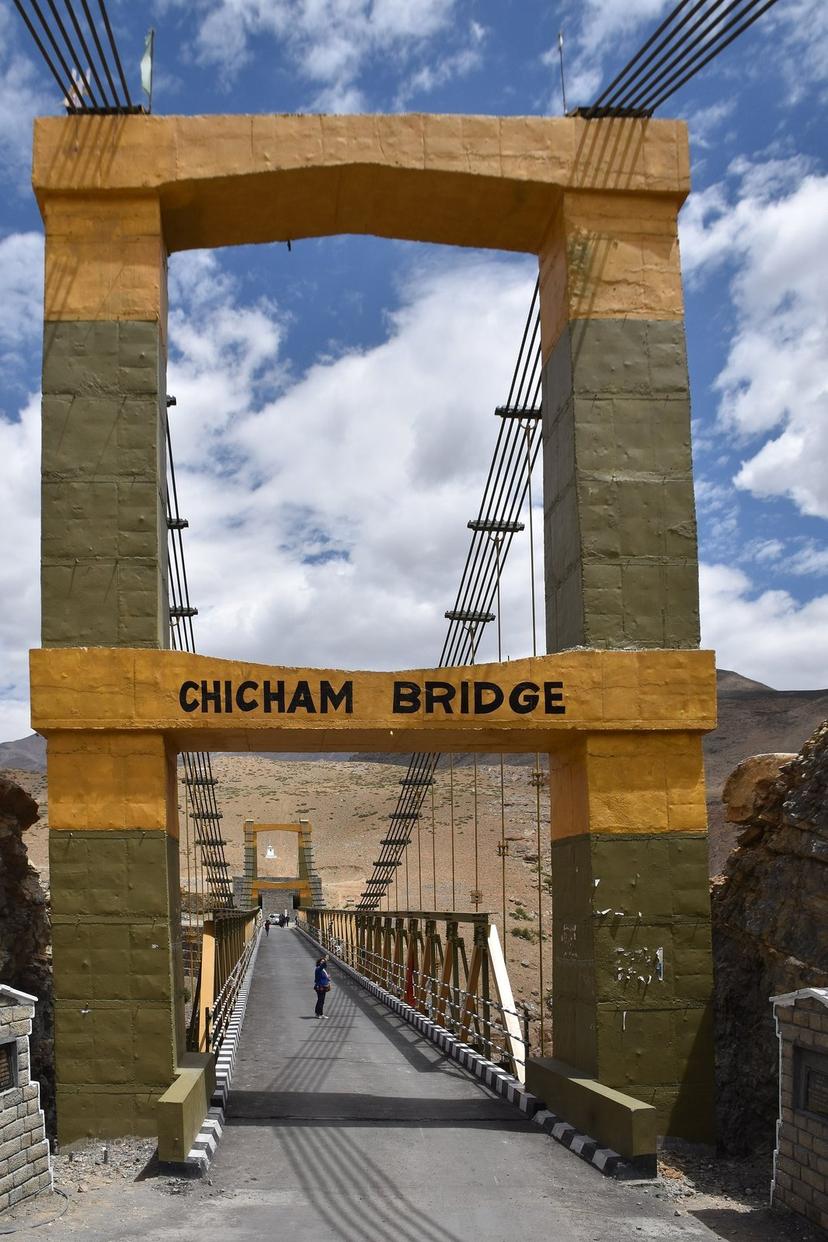 Yellow painted concrete archway entrance to Chicham Bridge in Spiti, with the steel suspension bridge deck stretching back behind it across a deep gorge. A single figure walking across in the distance