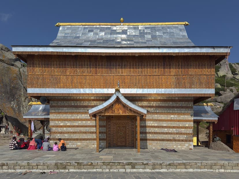 Front of a Himachali kath-kuni temple showing alternating horizontal courses of timber and dry stone under a silver slate roof, with a carved wooden door