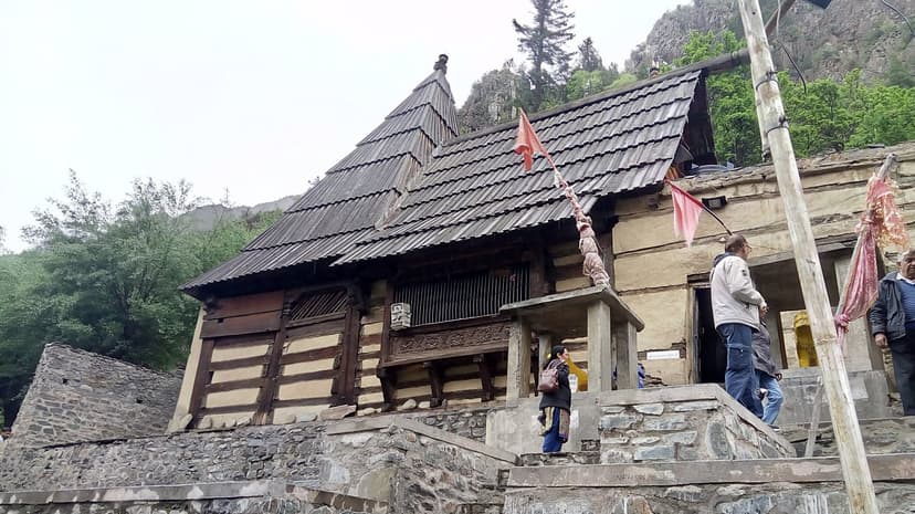 Mrikula Devi Temple at Udaipur in Lahaul, a pagoda-style wooden temple with a weathered slate roof, saffron flags, and carved wood panels visible in the dark inner facade