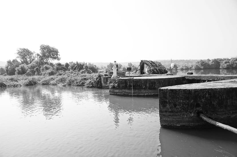 A traditional stone-and-earth khazan sluice-gate embankment on a tidal river at Moira, Goa, with a small shrine and thatched shelter on the bund, in black and white.