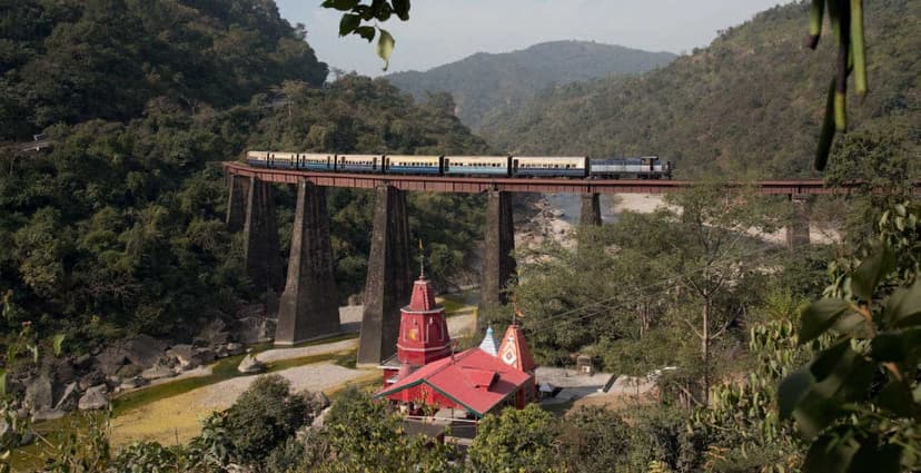 A narrow-gauge Kangra Valley Railway train rolling across the Bathu viaduct, with a small red-roofed Hindu shrine in the gorge below