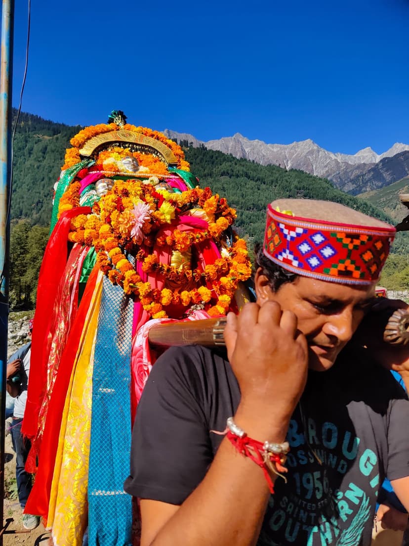 A Kullu devotee in an embroidered red-and-silver Kulluvi topi adjusting his earring with a marigold-draped deity palanquin behind him and snow peaks in the distance