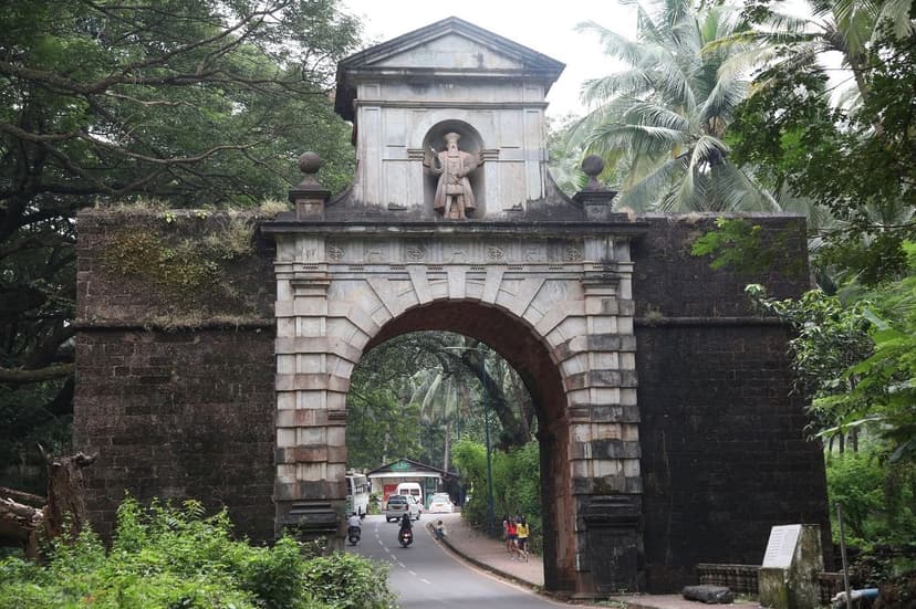 The laterite-and-stone Viceroy's Arch in Old Goa, a statue of Vasco da Gama in the upper niche, a road with a scooter passing under it, palms behind