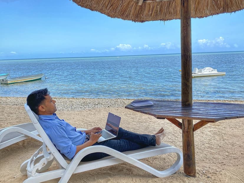 A man reclining on a beach sun-lounger under a thatched umbrella, working on a laptop by a turquoise sea