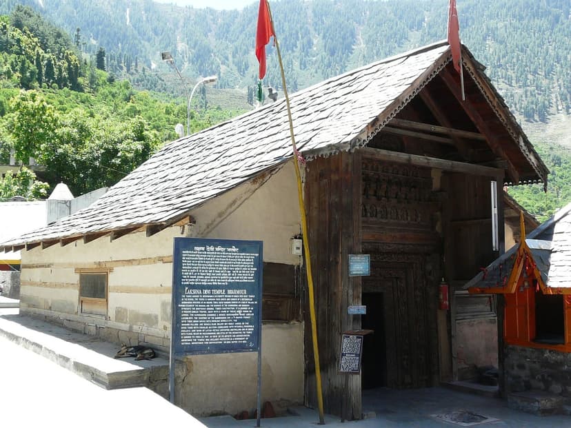 Exterior of the Lakshana Devi Temple at Bharmour — slate-roofed wooden structure with intricately carved doorway, ASI signboard, and red flags, with forested hills rising behind